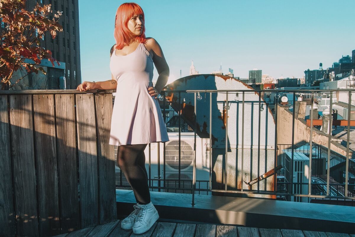 A pink haired woman wearing a lavender Halara dress, stands on a rooftop against a clear blue sky, with a hand on her hip.