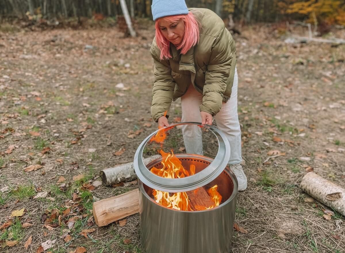 A woman i a green puffy coat with pink hair places the metal rim on a Solo Stove as bright orange flames roar inside.