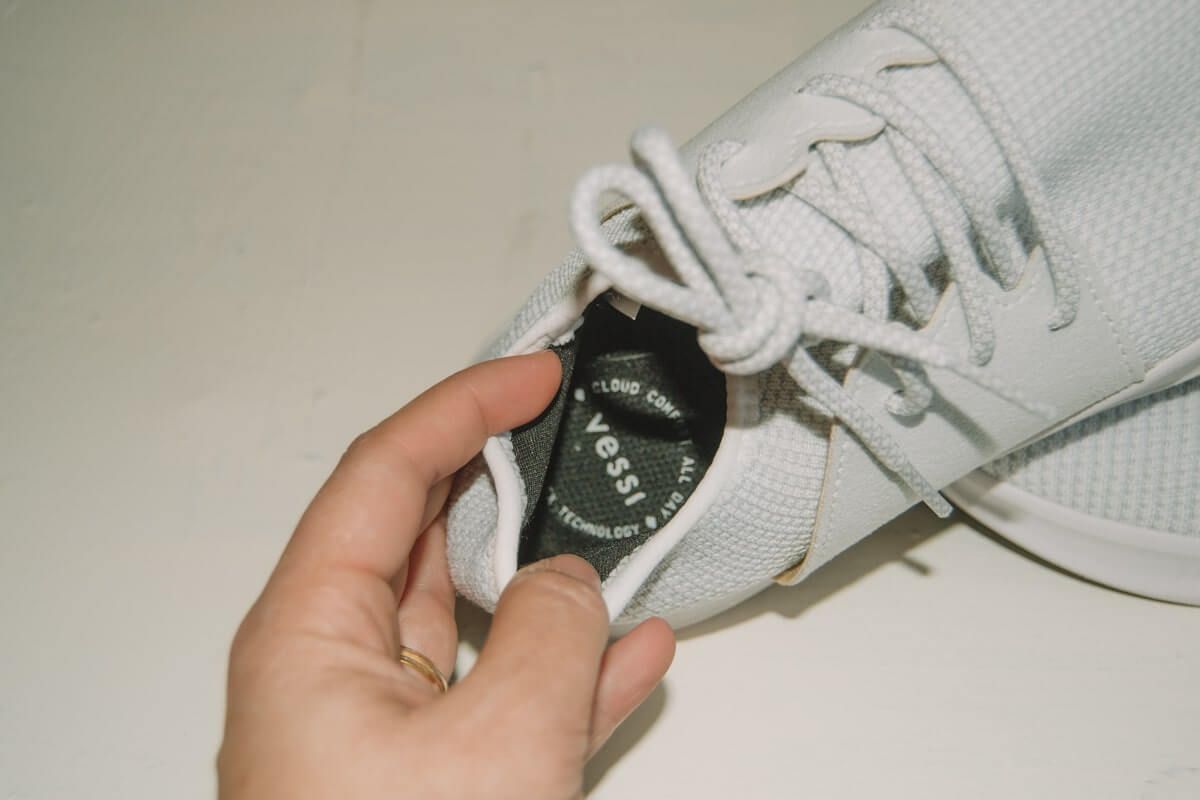 A hand holding a white Vessi sneaker open to show the logo on the insole against a white background.