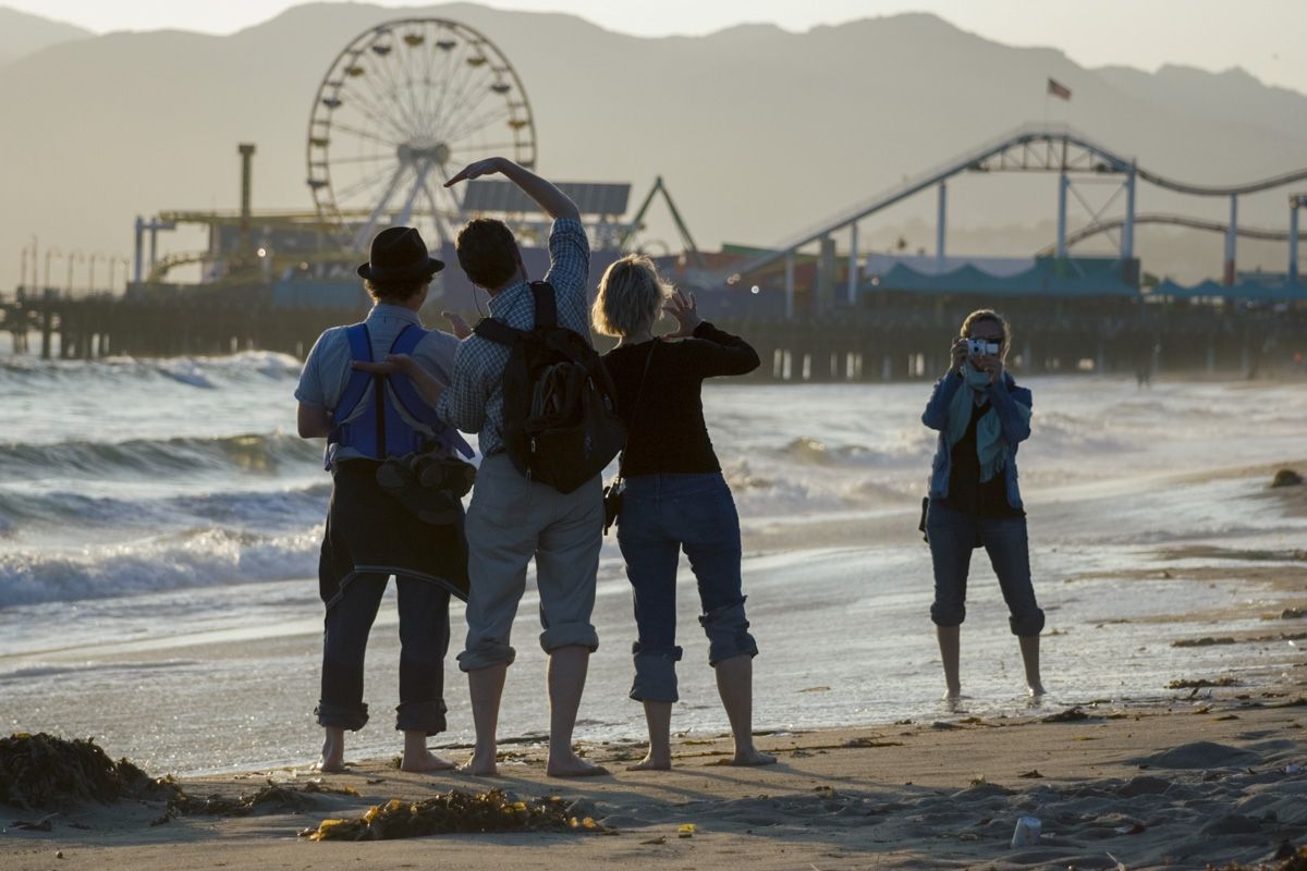Three people on a beach with their backs turned, facing a pier with a Ferris wheel and roller coaster, while one person takes a photo of them.