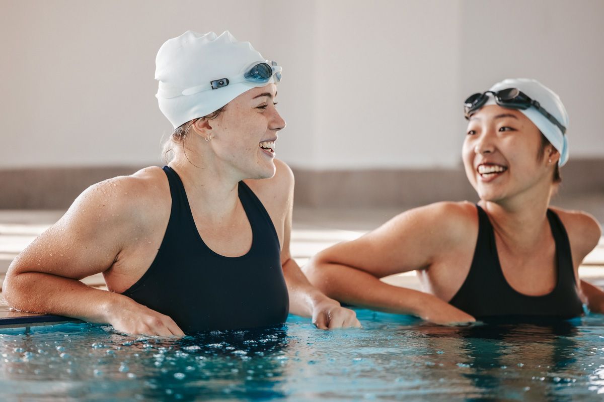 Two female swimmers in black bathing suits wearing examples of some of the best gifts for swimmers-- swim caps and goggles-- laughing togehter as they lean against the side of an indoor pool.