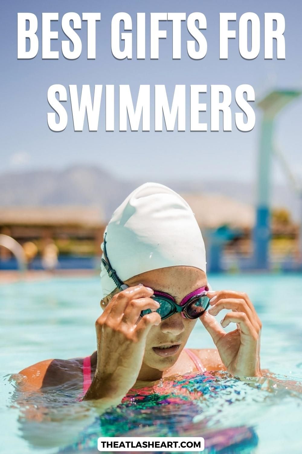 A young female swimmer in a white swimcap pokes here head out of the pool to adjust her goggles on a sunny day, with the background in soft focus, and the text overlay, "Best Gifts for Swimmers."
