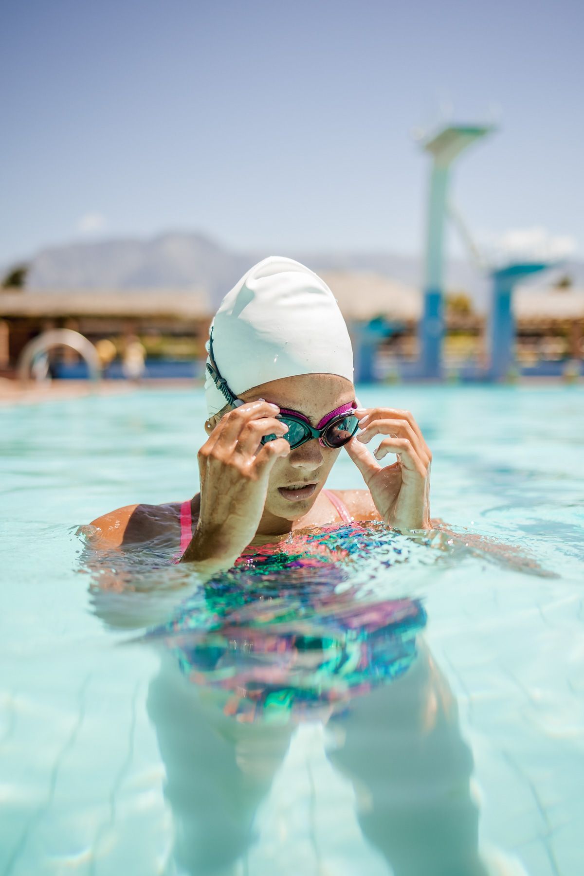 A young female swimmer in a white swimcap pokes here head out of the pool to adjust her goggles on a sunny day, with the background in soft focus.