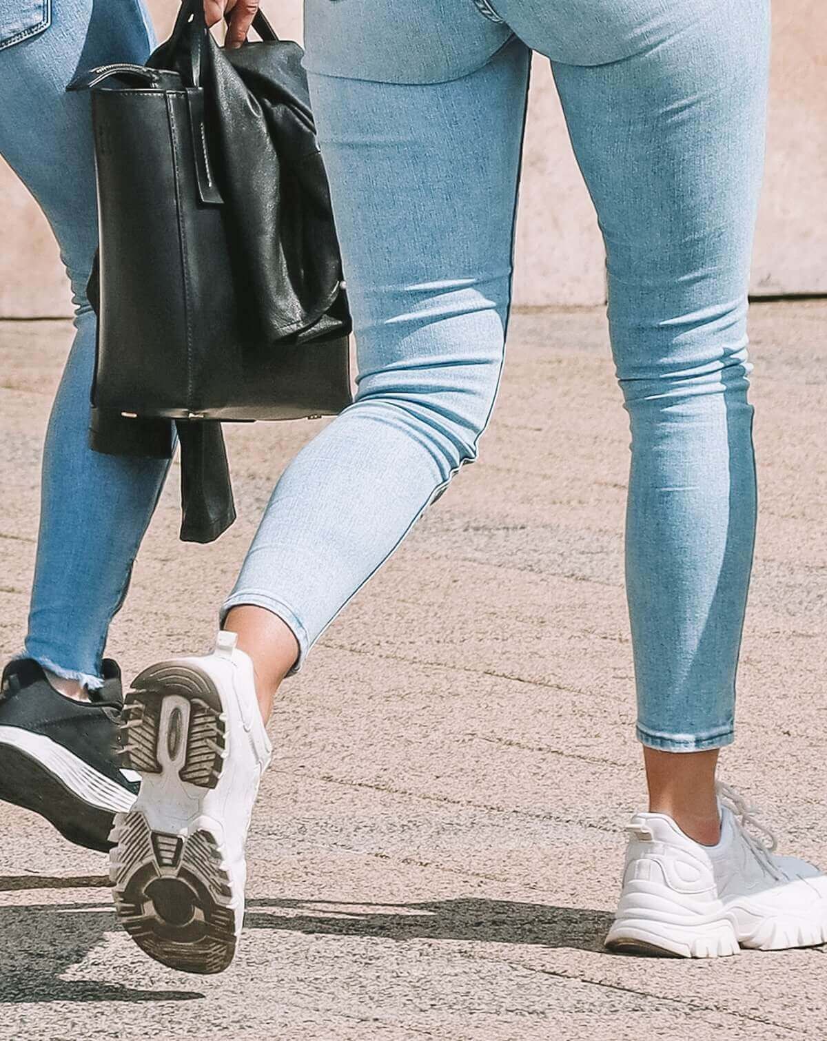 Two sets of women's legs wearing jeans and comfortable walking shoes, some of the best shoes for Disneyland, strolling on a concrete sidewalk on a sunny day.