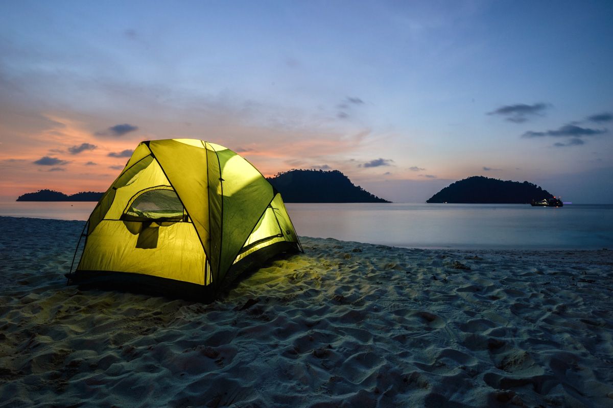 A yellow tent illuminated from the inside, set up on a sandy beach during a blue and pink twilight with a calm sea in the background.