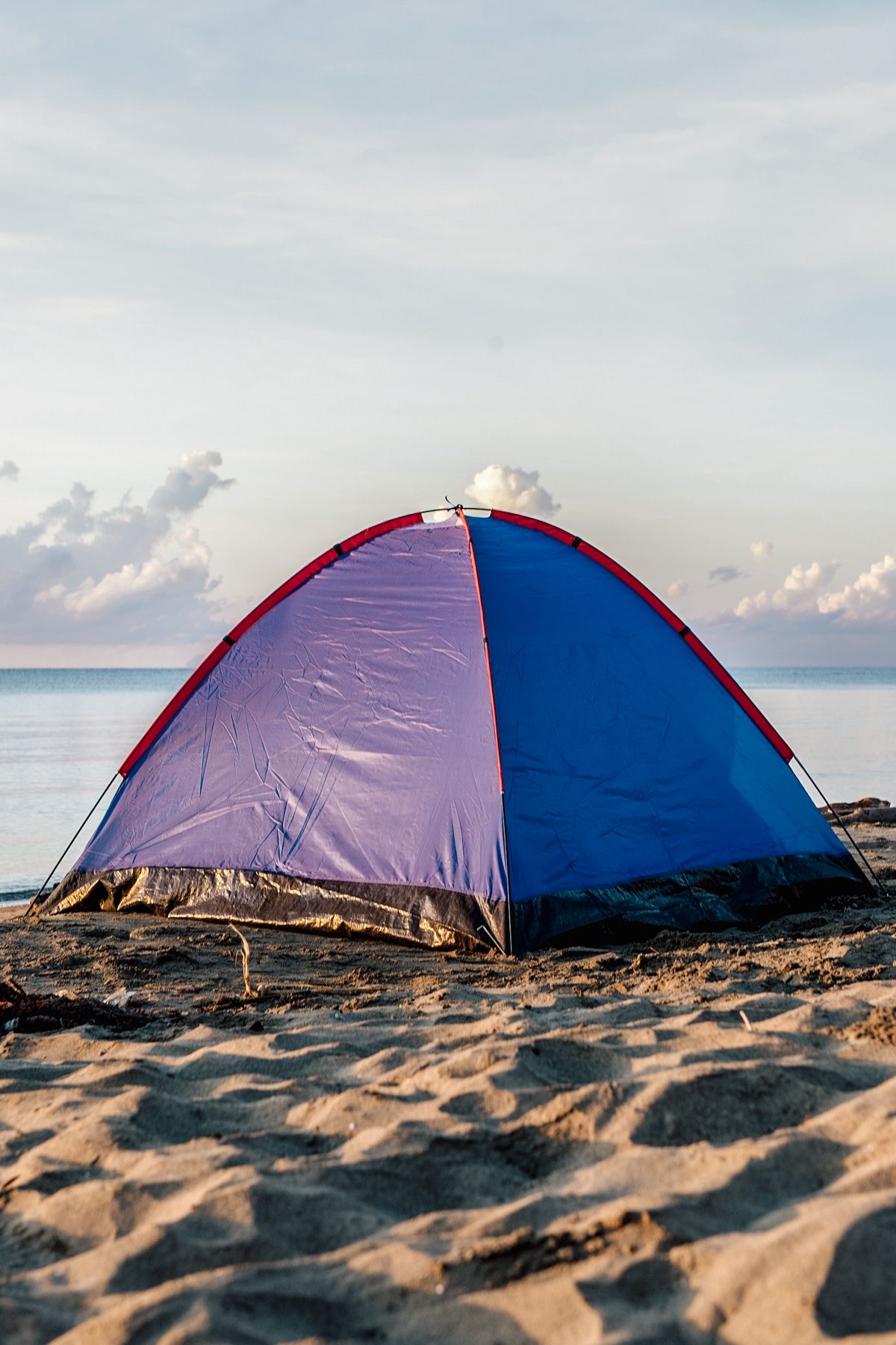 A blue and purple tent on a sandy beach with a calm sea and a partly cloudy sky in the background.