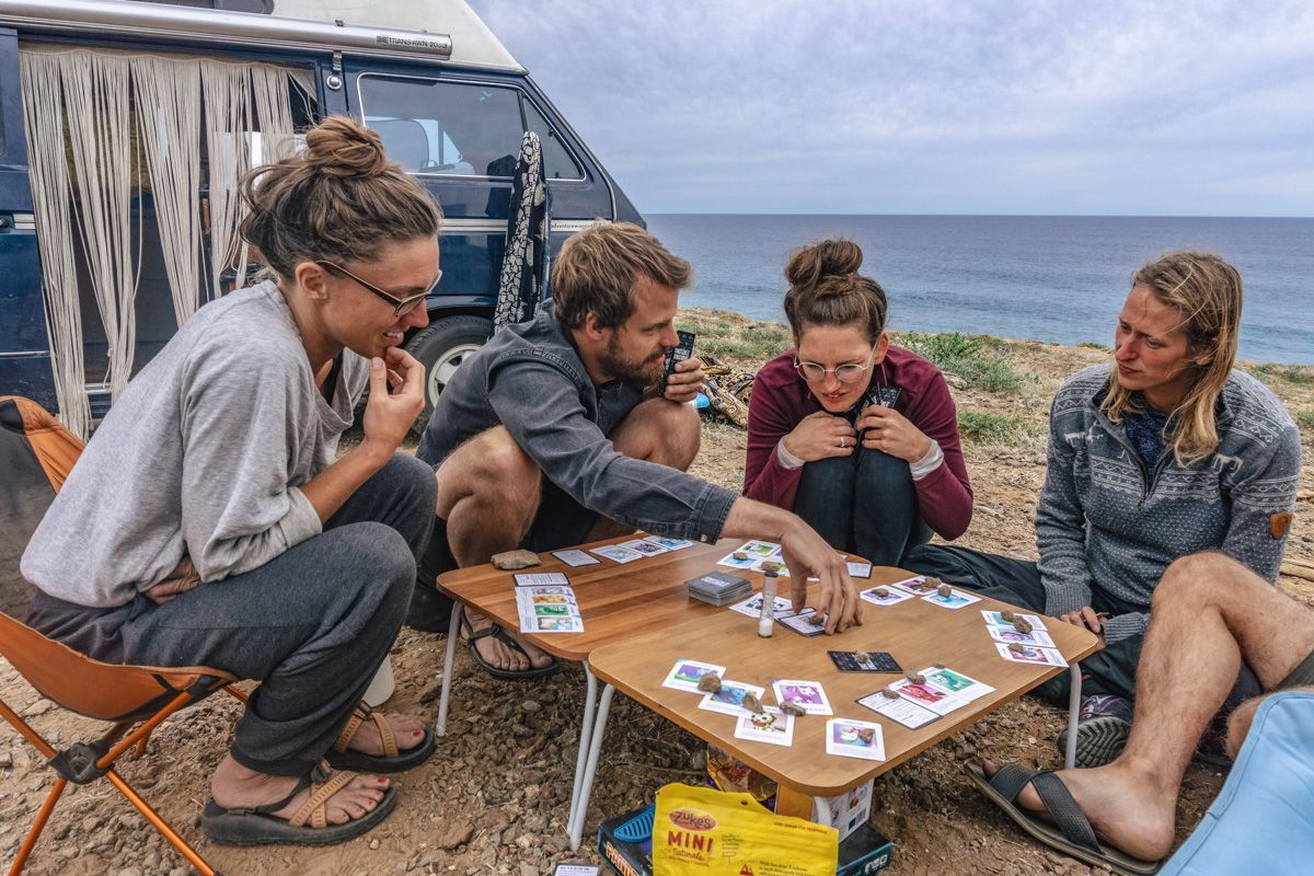 Card Games 
A group of five adults sitting around a knee-high table, playing cards, with a camper van and the ocean behind them.