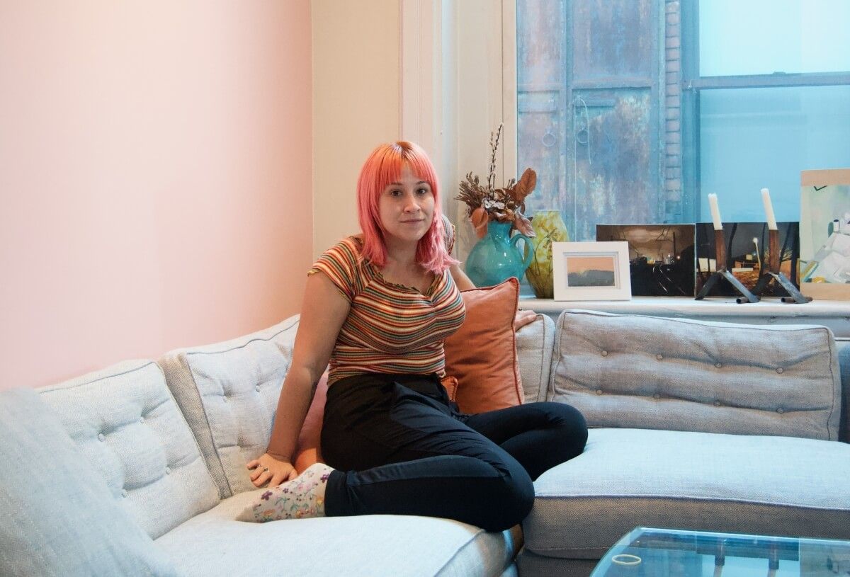 A pink-haired woman wearing a striped t-shirt and black pants sits with her legs folded under her on a grey-blue couch in front of a pink wall and a window.