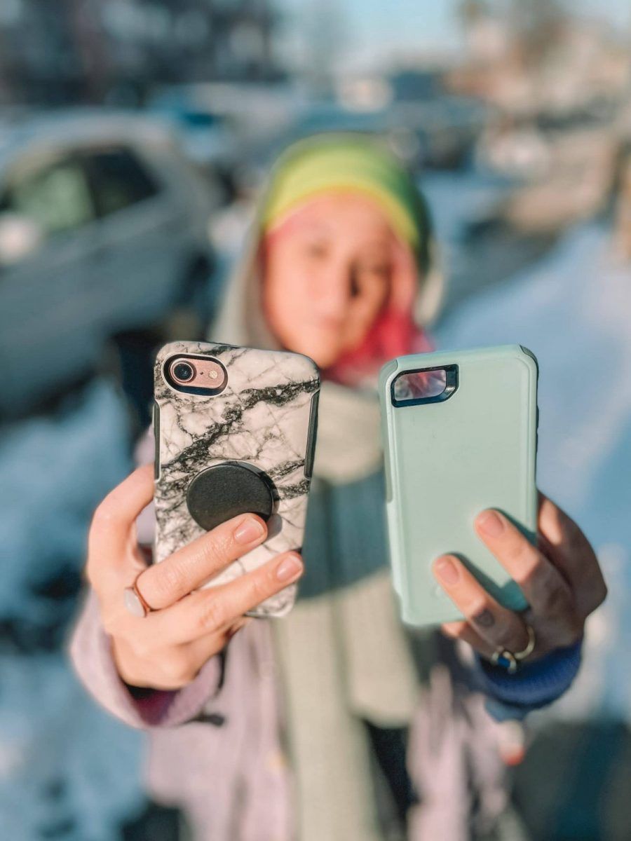 A pink-haired woman in a yellow hat in soft focus holds a white marble print phone case and a light teal one out in front of her, standing on a snowy city street.