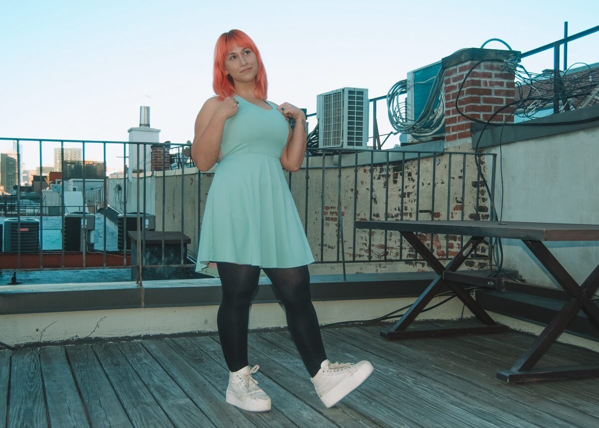 A pink haired woman stands on a rooftop, wearing a light green dress and white sneakers.