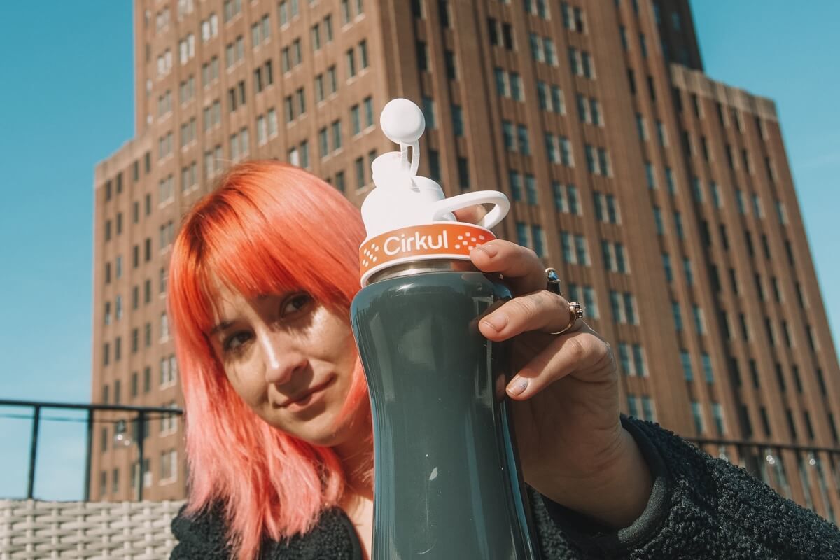 A woman with pink hair holding a Cirkul water bottle in front of her and eyeing it knowingly, with a blue sky and building in the background.