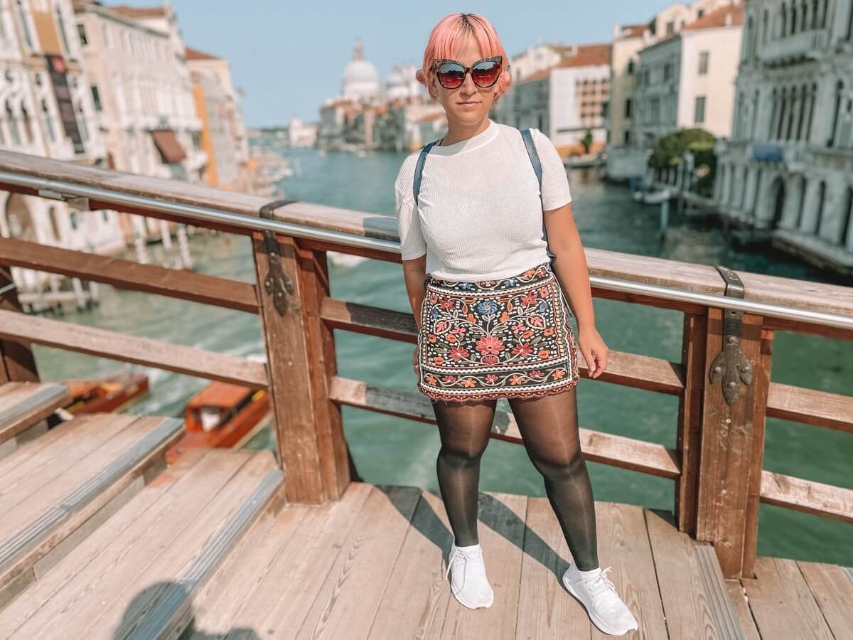 A woman with pink hair wearing sunglasses, a white t-shirt, an embroidered mini-skirt, black tights, and white Vessi shoes stands on a bridge in Venice.