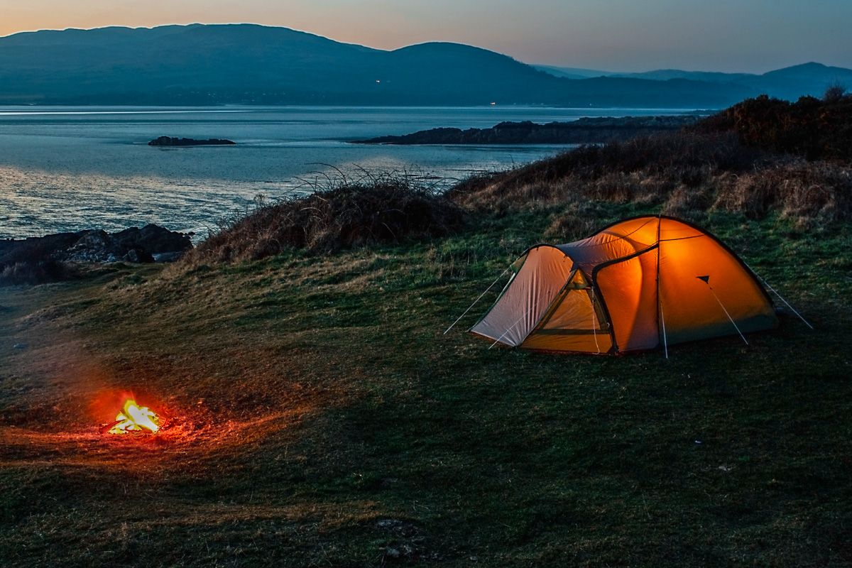 A glowing orange tent pitched on a grassy shore, illuminated by a nearby campfire, with a serene backdrop of the sea and silhouetted blue mountains during twilight.