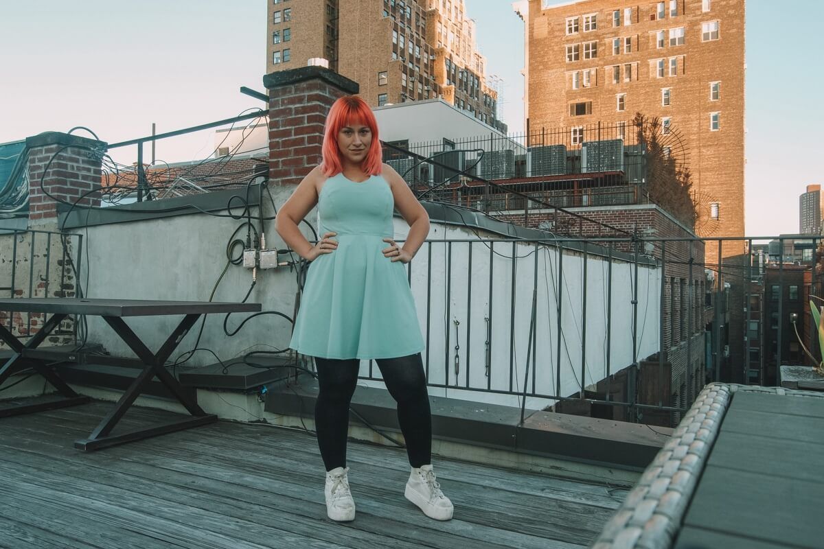 A pink haired woman stands with her hands on her hips in the shade on a rooftop, wearing a light green dress.