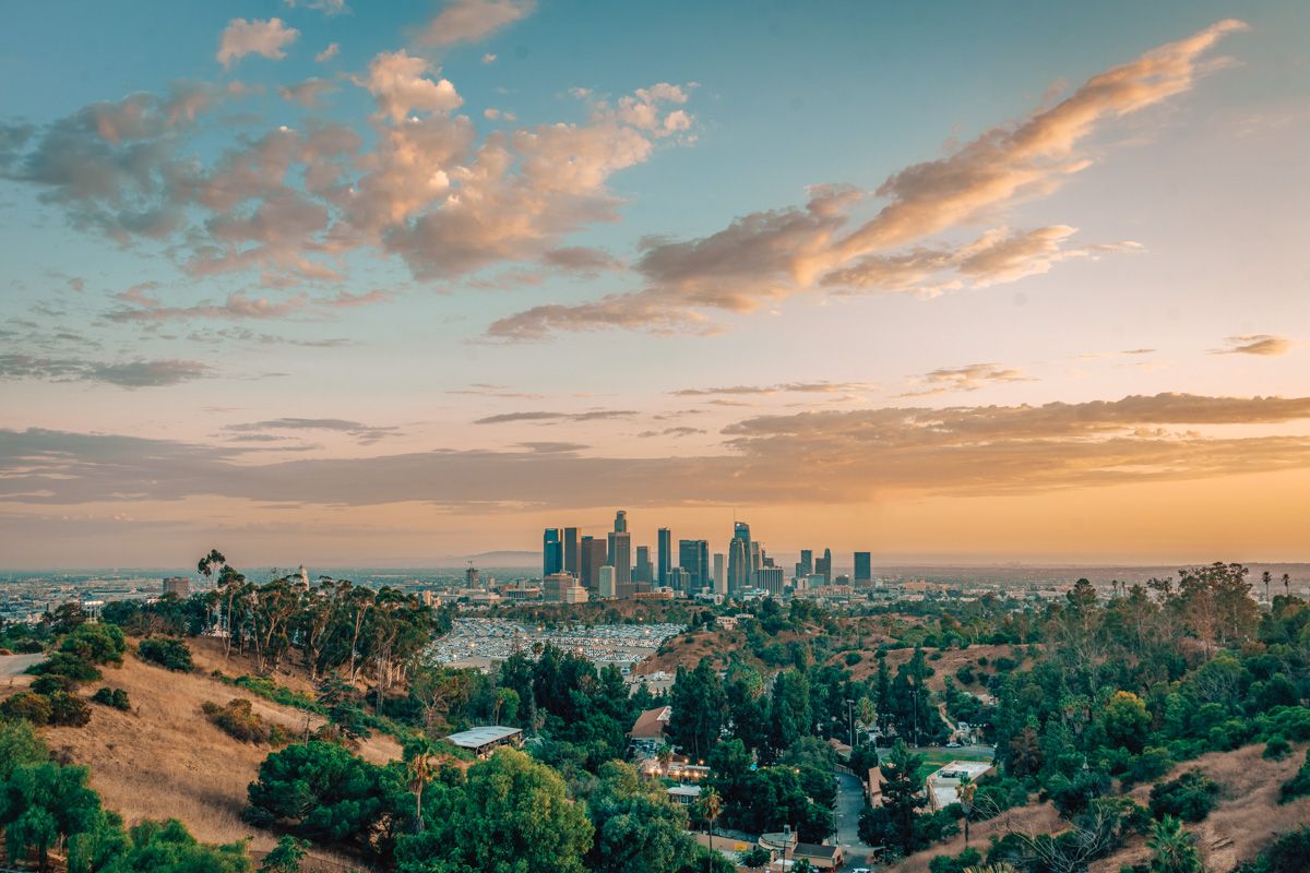 Golden hills dotted with everygreen trees, with Downtown LA visible beyond against a blue and yellow sunset.