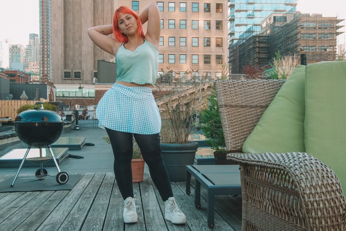 A pink haired woman wearing a blue tank top and a checkered blue and white skirt, stands on a rooftop against a clear blue sky, with a hand on her hip.
