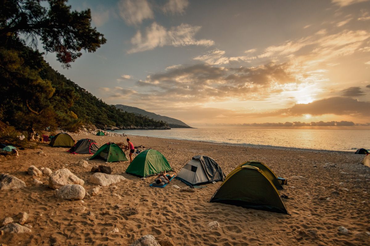 A sandy beach at sunset with six dome tents set up in a line along the beach, near a forested hill.