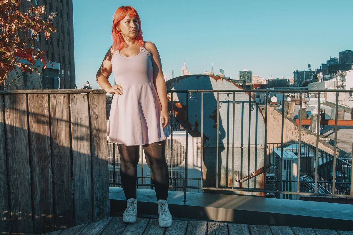 The author of this Halara review, a pink haired woman wearing a lavender Halara dress, stands on a rooftop against a clear blue sky.