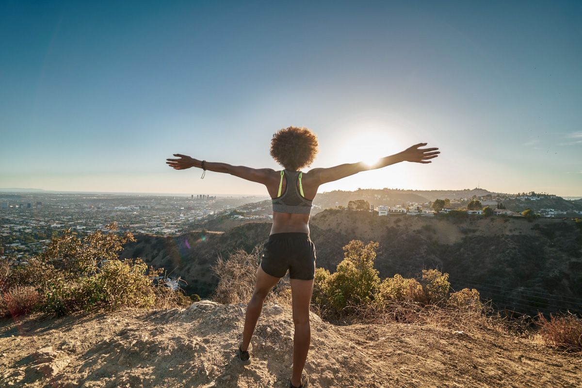 A woman in bike shorts and a sports bra seen from behind after completing one of easy hikes in Los Angeles, standing at a lookout point with her arms outstretched.