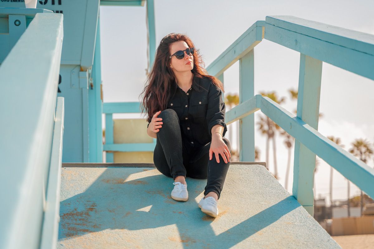A dark-haired woman dressed in black and wearing sunglasses sitting on a blue lifeguard tower, with palm trees in the background.