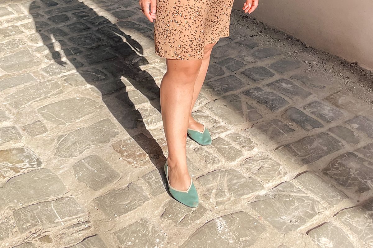 A pair of feet wearing a pair of teal Vivaia barefoot dress shoes for women and standing on a cobblestone street.
