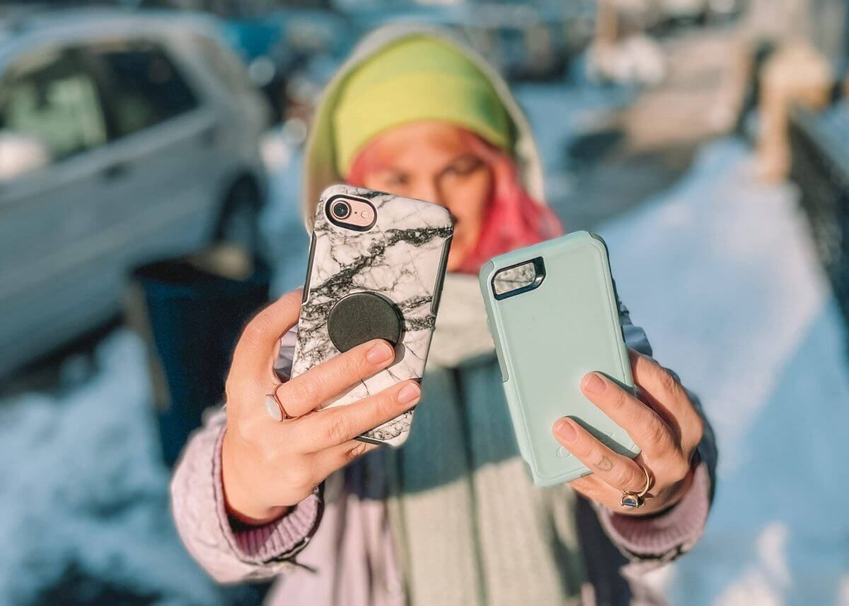 A pink-haired woman in a yellow hat compares the Otterbox symmetry vs commuter by holding phone cases out in front of her, standing on a snowy city street.