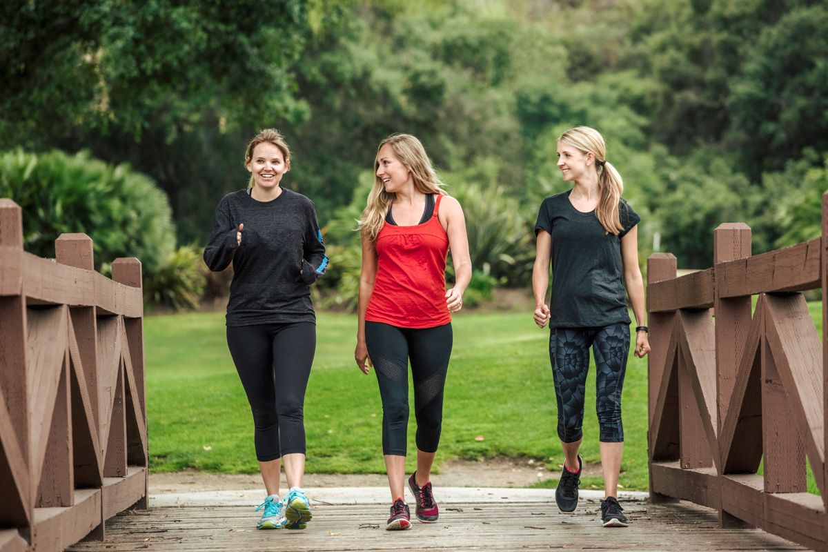 Three blond women in red and black athletic wear walking on a wooden bridge in a lush park setting.