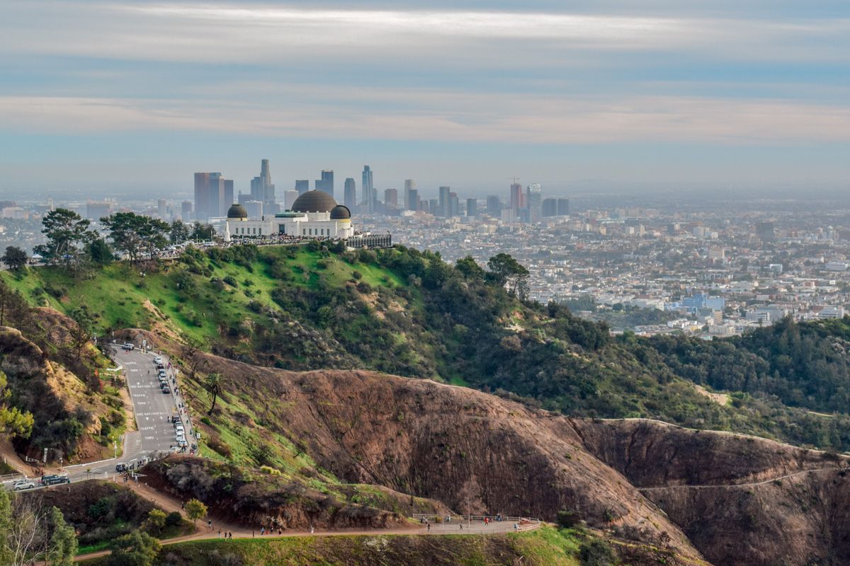 A view of Griffith Observatory on the top of a green hill, with the sprawl of Los Angeles and a hazy sky in the background.
