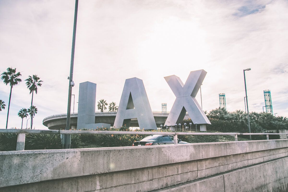 The entry sign for Los Angeles International Airport (LAX), seen from the freeway, with palm trees silhouetted against a partly cloudy sky.