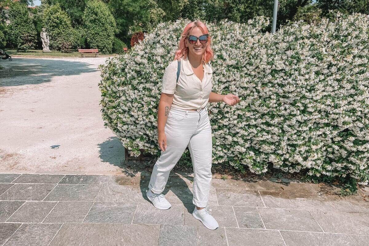 A woman with pink hair wearing sunglasses, a white blouse, white jeans, and white Vessi shoes in front of a bush covering in white flowers, laughing a the camera.