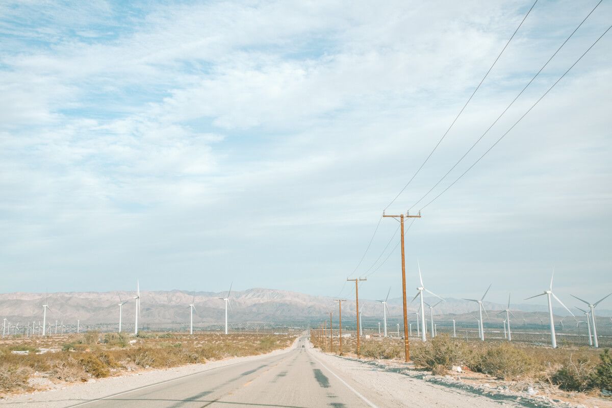 An open road in a California desert landscape dotted with windmills, and hazy, dry hills visible in the distance.