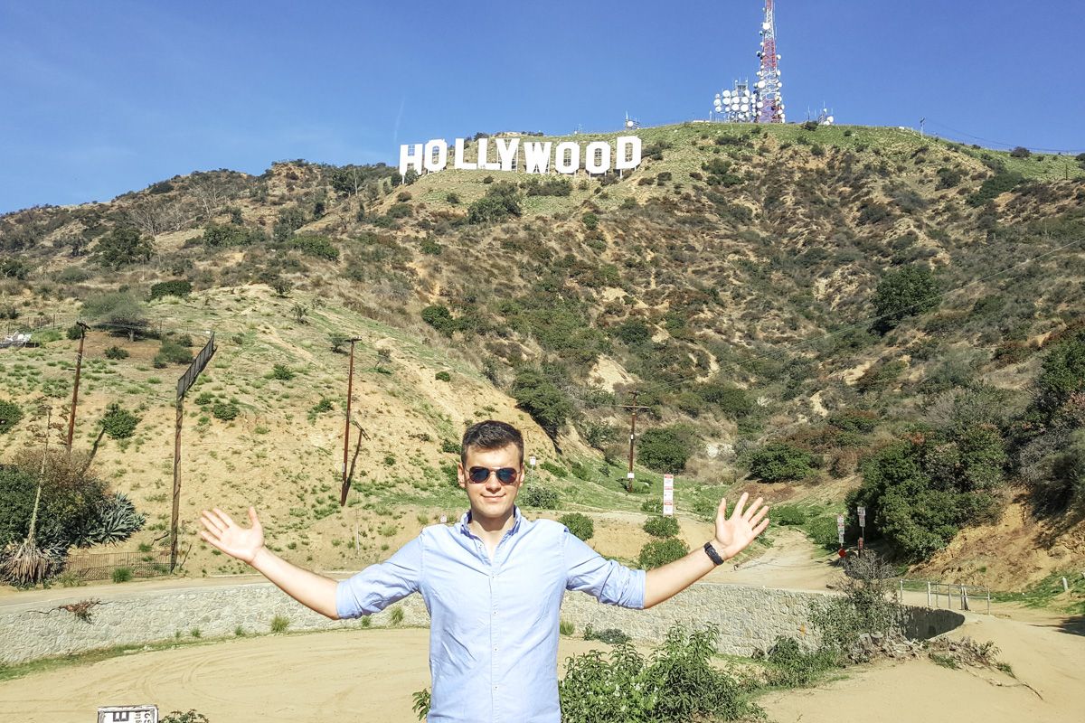 A man with sunglasses and a blue shirt stands in the foreground with arms outstretched, the iconic Hollywood sign on a hill in the background.