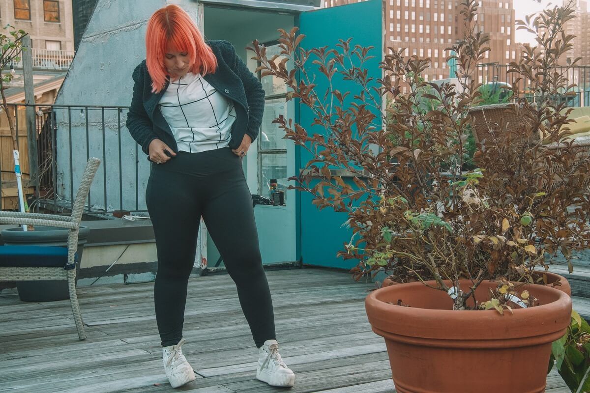 A pink-haired woman wearing the Patitoff pet hair resistant high-waisted crossover plain leggings and a black and white grid-print shirt stands on a roof and looks down at her feet.