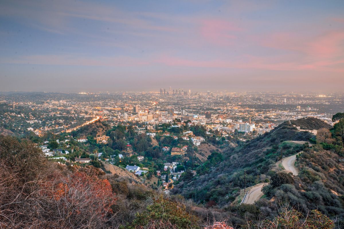 A view from the top of a hill, looking out at Runyon Canyon and a Los Angeles cityscape at early sunset.