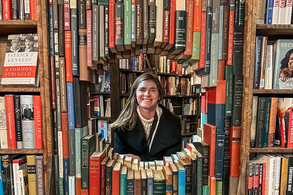 A smiling woman in a black coat and white sweater stands amidst towering bookshelves filled with various titles, with a unique archway of stacked books in front of her.