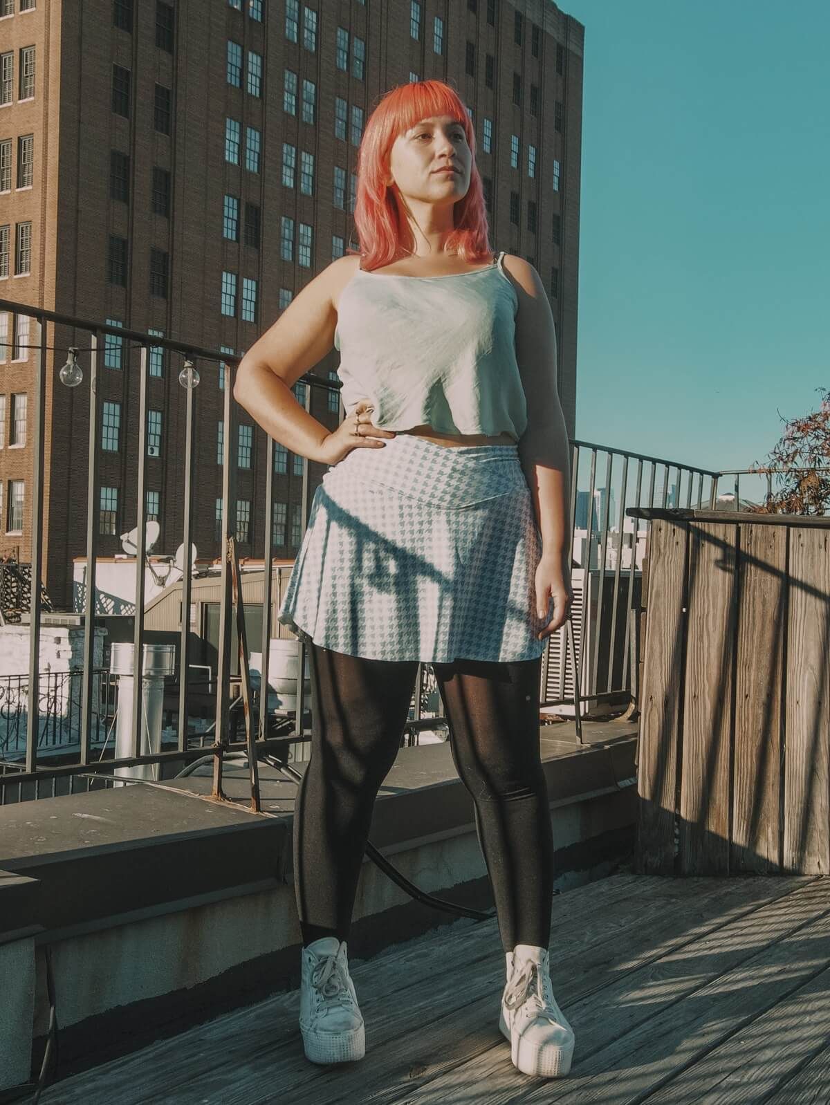 A pink haired woman wearing a blue tank top and a checkered blue and white skirt, stands on a rooftop against a clear blue sky, with a hand on her hip.