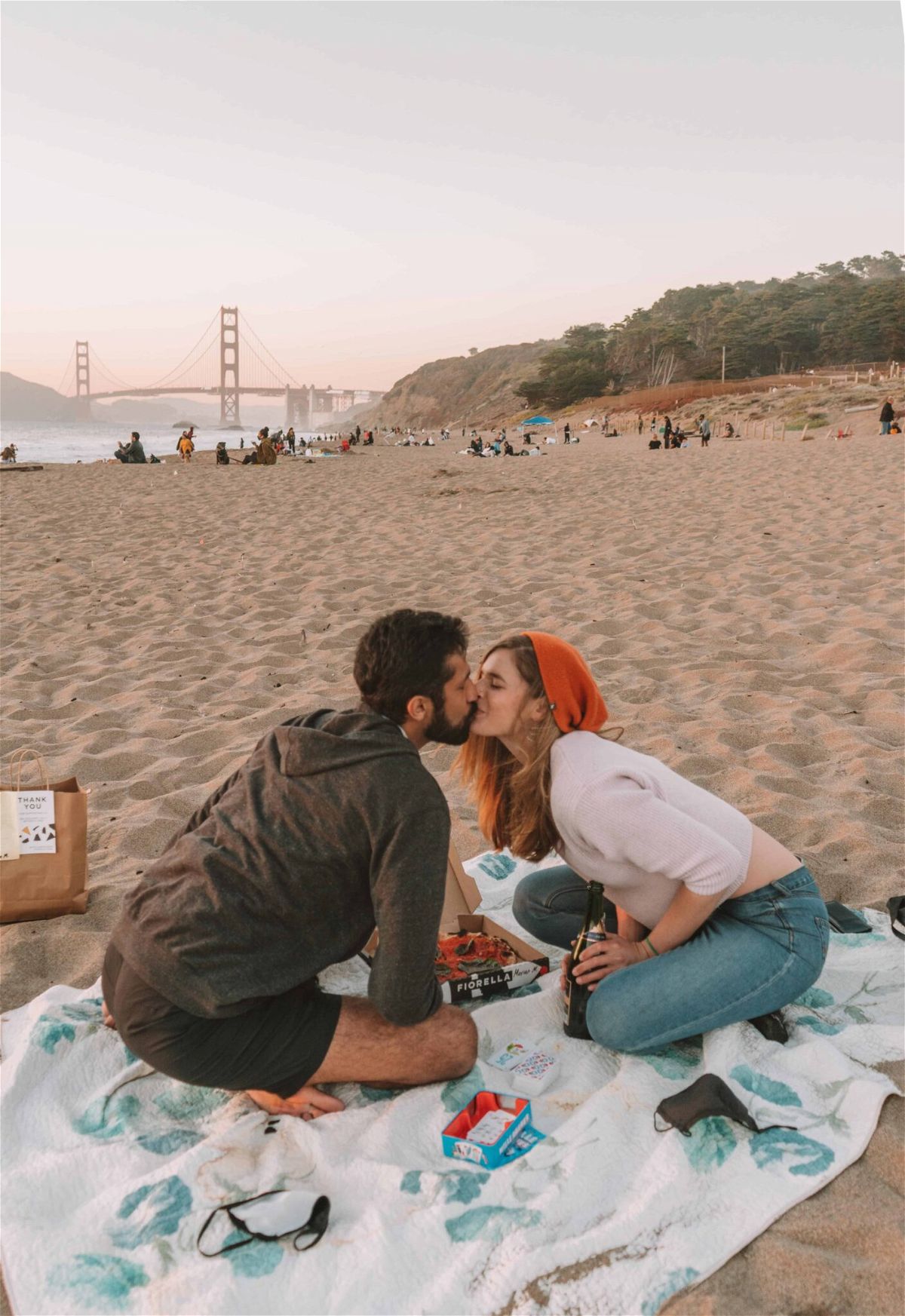 A man and woman share a kiss over a pizza and a bottle of champagne, sitting on a picnic blanket on the beach with the Golden Gate Bridge in the background.