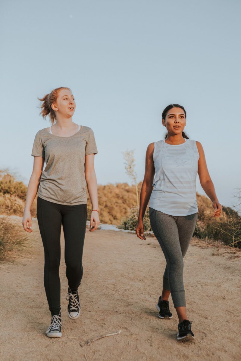 Two women in exercise clothes walking side-by-side along a wide, easy trail with a dim, cloudless sky behind them.