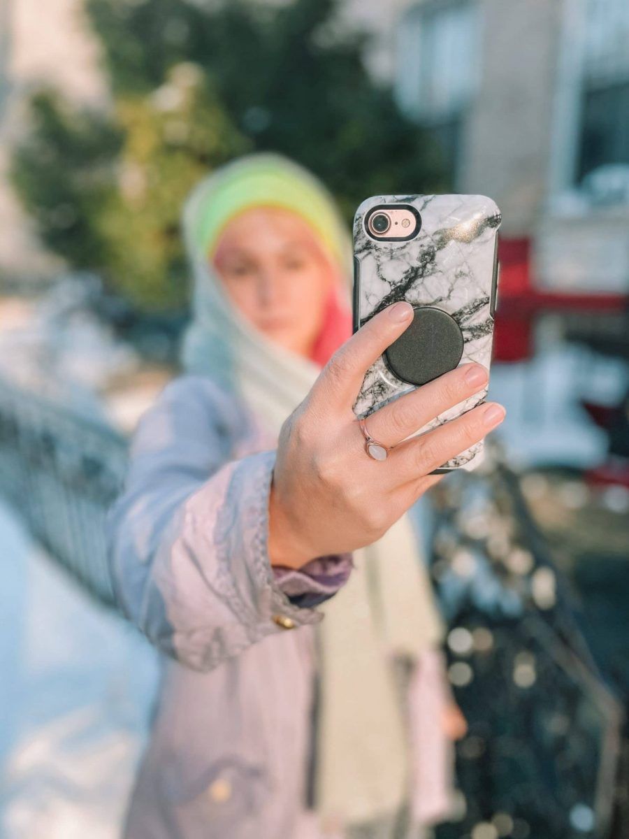 A pink-haired woman in a yellow hat in soft focus holds a white marble print phone case out in front of her, standing on a snowy city street.