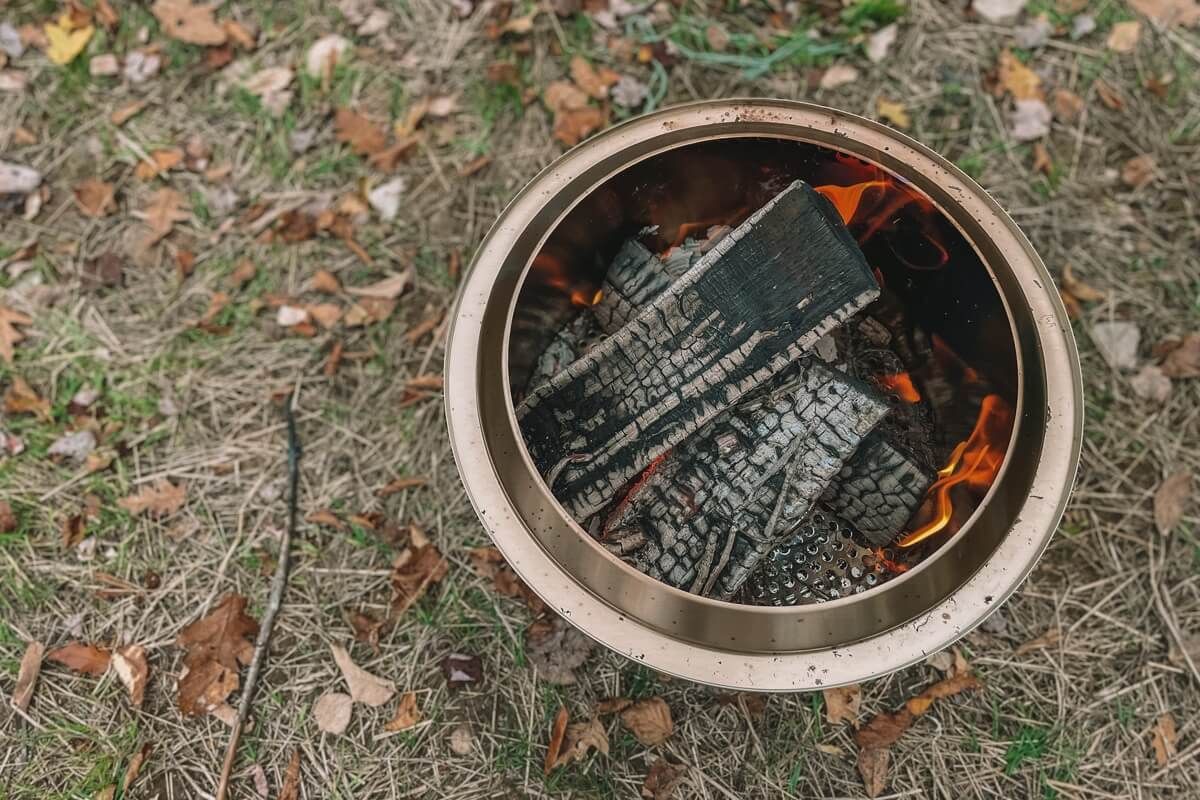 Several pieces of charred firewood and flames burning inside of a Solo Stove sitting on the ground outdoors.
