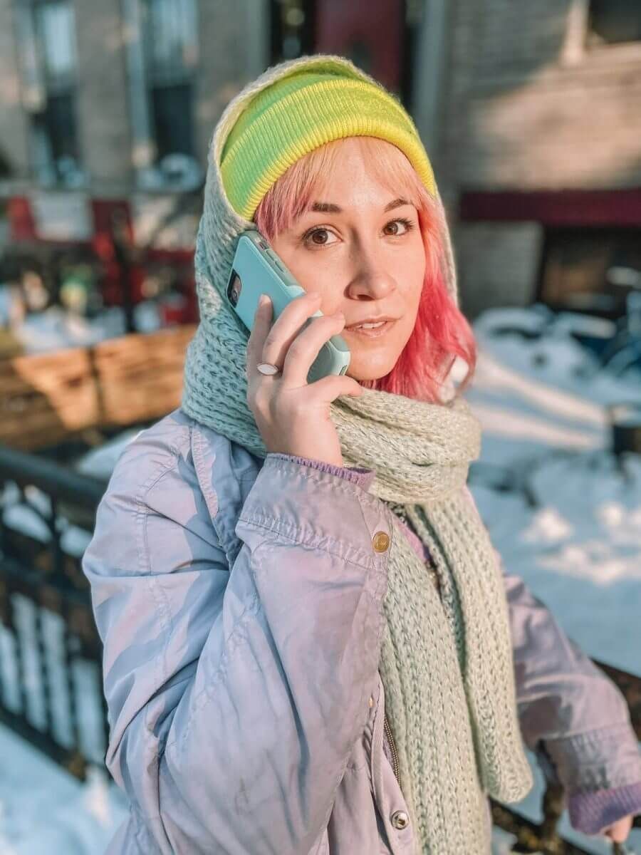 A pink-haired woman in a yellow hat in soft focus holds a phone in. alight teal case up to her ear, standing on a snowy city street.