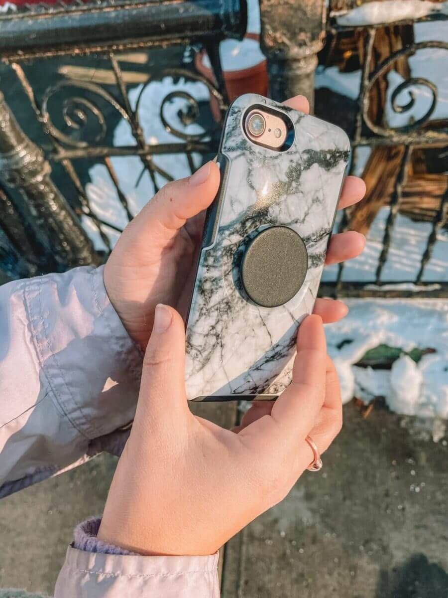 A pair of hands holding a white marble print Otterbox Symmetry phone case with a black pop socket in profile with a snowy city street in the background.