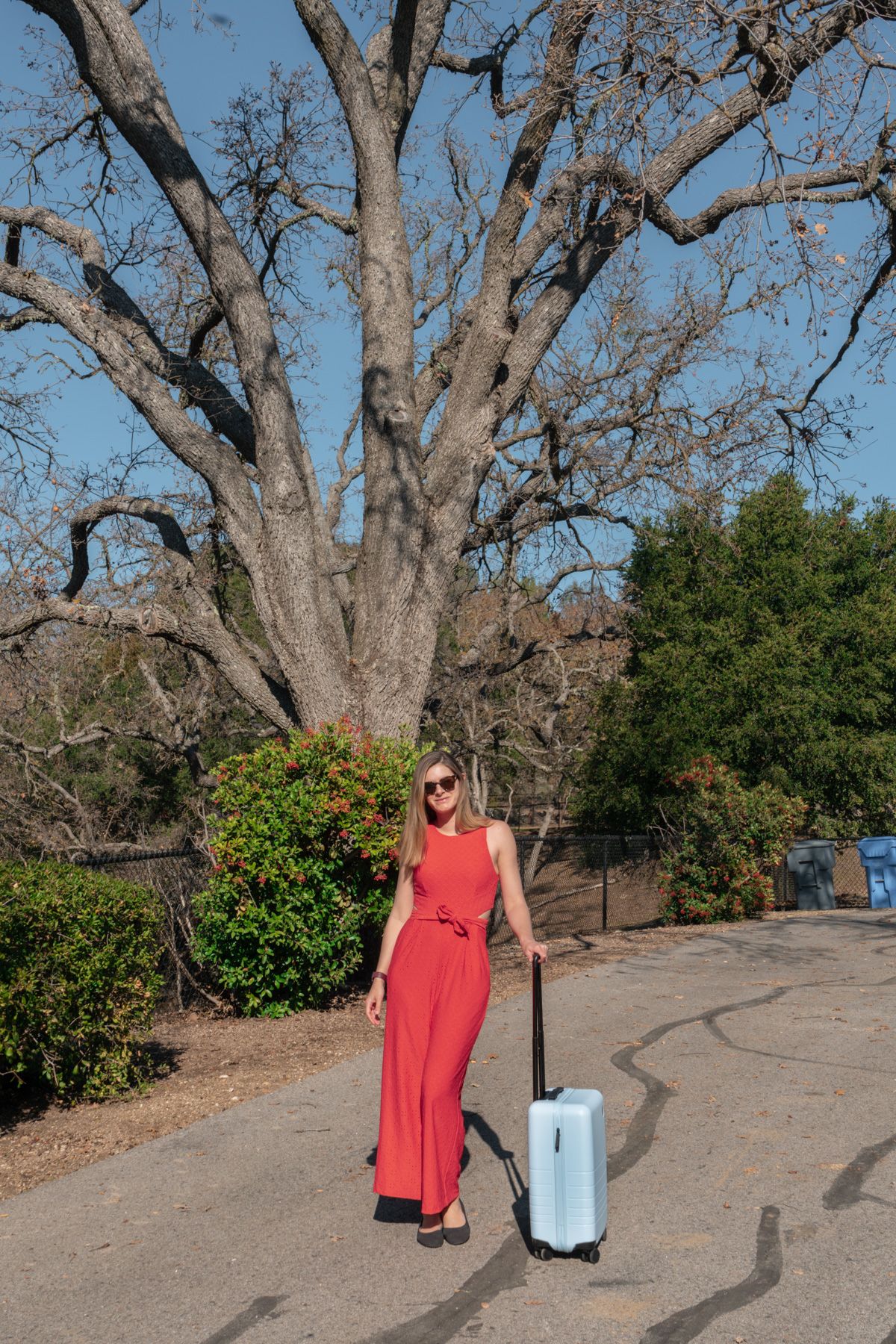 Woman in red dress and sunglasses holding a light blue hardshell suitcase outdoors with trees and a clear blue sky in the background.