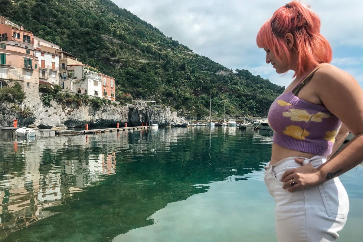A pink-haired woman wearing a purple, flower-print, knit crop top and white jeans stands in profile with a harbor and green hill in the background.
