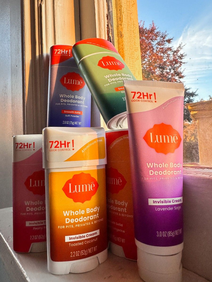 A stack of Lume deodorants in various scents, stacked on a windowsill, with a blue sky and autumn leaves visible through the pane.