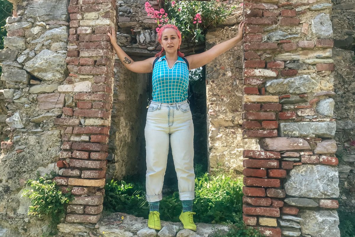 A woman with pink hair in a blue and white checkered top, light-wash jeans, and neon green shoes stands in a crumbling stone doorway of an ancient ruin.
