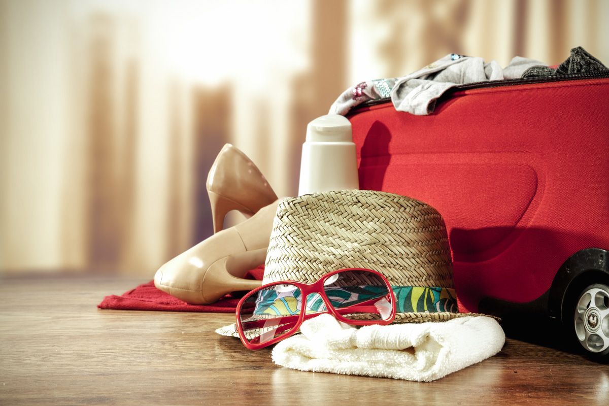 A pair of sunglasses, a hat, a towel, and other travel items laid out next to a red suitcase.