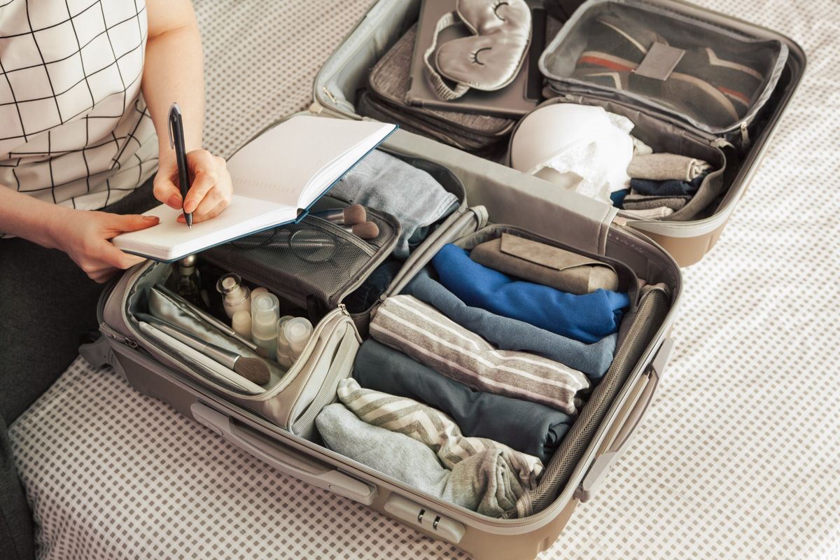 A view from above of a woman's hands writing in a blank notebook balanced against an open suitcase filled with clear packing cubes, which is sitting on a bed.