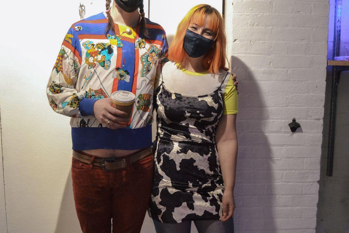 A woman with orange hair smiles beneath a face mask while wearing a cow-print velvet minidress, her arm around a tall man in brightly-colored clothing.