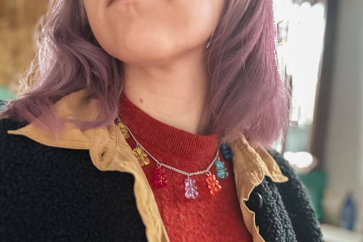 A close-up of a purple-haired woman's neck wearing a Cartoon Bear Charm Necklace and an orange sweater.